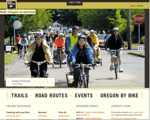 A group of cyclists including men and women wearing helmets ride together on a street during a biking event with trees and a stop sign visible in the background The website menu promotes trails events and resources
